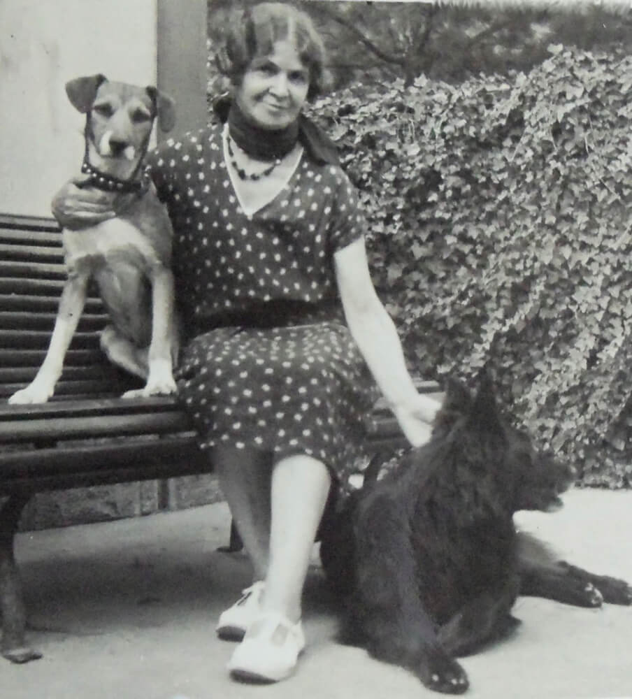 Photographie représentant Jeanne L'Enfant dans un jardin, assise sur un banc, en compagnie de ses deux chiens.